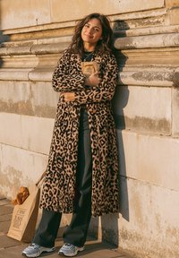 Žena oblečená v kabáte s leopardím vzorom stojí pri kamennej stene, drží zabalenú bagetu a vedľa nej na zemi je taška s chlebom.