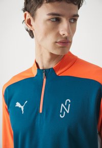 Puma maglia da uomo con zip, caratterizzata da un corpo teal con accenti arancioni sulle spalle, tessuto testurizzato e logo stampato sul petto.