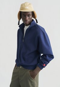 Φούτερ με φερμουάρ σε ναυτικό μπλε χρώμα, με ψηλό γιακά, χαλαρή γραμμή και ριμπ μανσέτες. Συνδυάζεται με παντελόνι σε ελαιόχρωμο με πλαϊνό λογότυπο.