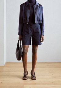 Laivastonsininen lyhyt takki vetoketjulla, yhdistettynä laivastonsinisiin shortseihin. Ruskea käsilaukku ja remmisandaalit täydentävät asun. Taustalla puulattia.