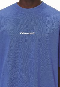 Modré bavlněné tričko s kulatým výstřihem a krátkými rukávy, na přední straně se bílým logem "PEGADOR". Hladká textura.