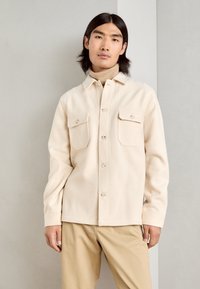 Beige textured shirt jacket with a button-up design, two chest pockets, and a classic collar, worn over a light turtleneck and neutral pants.