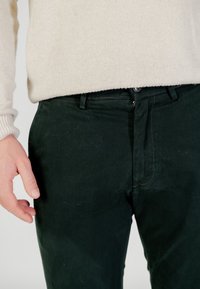 Pantalones de algodón verde oscuro con una textura suave, cierre de cremallera y botón, con detalles de costura sutiles y un ajuste entallado.