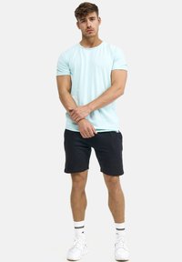 Light blue crew-neck t-shirt made of soft fabric, paired with black shorts. Model stands with arms crossed, wearing white sneakers with accents.