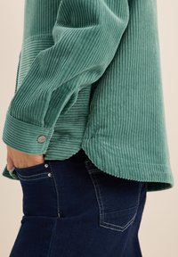 Henkilö, joka on pukeutunut teal-väriseen, ribbeillä varustettuun, corduroy-paitaan, jossa hihansuut on napitettuna, ja käsi tummansinisissä farkuissa olevan taskun sisällä.