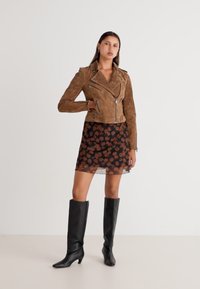 Jachetă de biker din suede maro peste o rochie neagră din material transparent cu flori, asortată cu bocanci negri ajustați până la genunchi. Modelul pozează pe un fundal deschis.