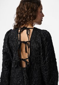 Crna haljina teksture sa napuhanim dugim rukavima i otvorenim leđima, sa trakama za podešavanje. Tkanina ima suptilan uzorak.