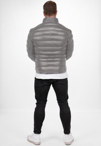 Grijze pufferjack met horizontale quilts, hoge kraag en een gladde textuur. Gecombineerd met zwarte spijkerbroeken en witte sneakers.