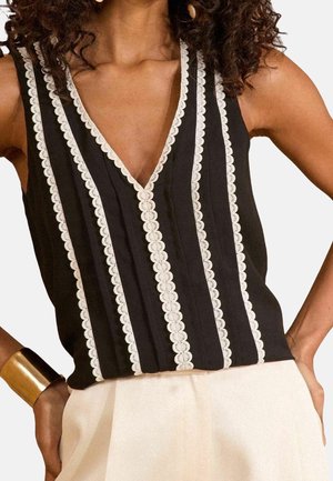 Mouwloze zwarte top met diepe V-hals en verticale witte scalloped kantstroken, gecombineerd met witte broek en een gouden armband.