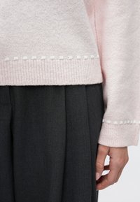 Ανοιχτό ροζ πλεκτό πουλόβερ με λευκές λεπτομέρειες στο κάτω μέρος, σε συνδυασμό με σκούρο γκρι πλισέ παντελόνι, που δείχνει πλάγια όψη του μανικιού και του χεριού.