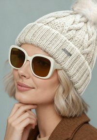 Knit beige beanie with a pom-pom, paired with large white sunglasses with brown lenses, highlighting a textured pattern on the hat.