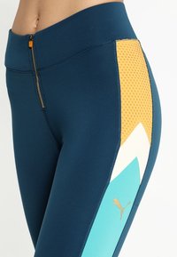 Höga tights i mörk teal med gul mesh och akvamarin färgdetaljer. Funktioner inkluderar en dragkedja bak och textil med struktur på sidorna.