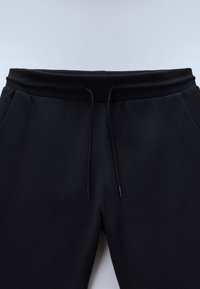 Mustad dressipüksid soonikuga elastse vöökohaga ja mustade paeltega, küljetaskutega, halli taustaga.