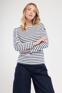 Kvinde med blond hår iført en langærmet hvid skjorte med sorte vandrette striber og mørkeblå bukser, stående med armene korslagte.