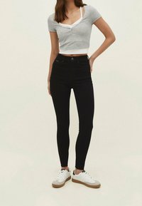 Jeans negros de cintura alta con un ajuste ajustado, combinados con un top corto gris claro con mangas cortas y detalles de botones. Zapatillas blancas completan el look.
