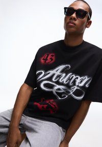 Fekete, túlméretezett póló fehér betűs felirattal, piros részletekkel és egy gyík grafikával. A modell fekete napszemüveget és világosszürke farmert visel.