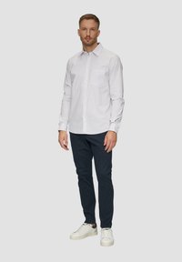Witte overeindknoopshirt met kleine blauwe stippen, gecombineerd met donkerblauwe broek en witte sneakers. Model staat voor een effen achtergrond.