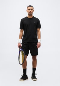 Άνδρας τενίστας όρθιος, ντυμένος με μαύρη στολή Lacoste, λευκά περιβραχιόνια, μαύρα αθλητικά παπούτσια, κρατώντας ρακέτα τένις στο δεξί του χέρι.