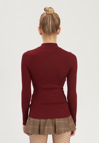 Donna con capelli biondi raccolti in un bun, che indossa un maglione aderente a coste color bordeaux, una gonna a pieghe a quadri marrone e calze velate nere, vista di spalle.