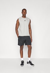 Haut de sport sans manches gris avec un logo noir, associé à un short noir et des baskets noires ; le modèle se tient devant un fond neutre.