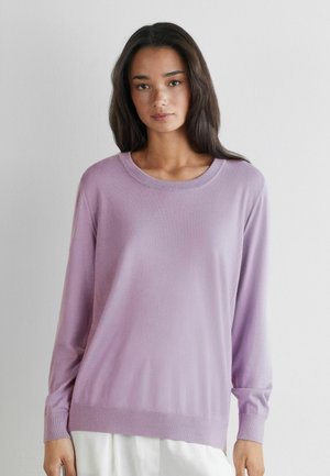 Kobieta z długimi ciemnymi włosami, ubrana w jasnofioletowy sweter z długim rękawem i białe spodnie, stojąca na tle jednolitej jasnej ściany.