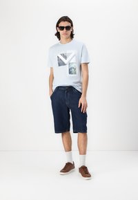 T-shirt en coton bleu clair avec des motifs graphiques, associé à un short en denim noir, des chaussettes blanches et des chaussures brunes ; mannequin portant des lunettes de soleil.