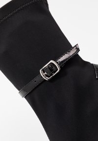 Botín de tela negra con una correa de charol negro brillante y detalle de hebilla plateada. Presenta una textura suave y bordes cosidos para mayor durabilidad.