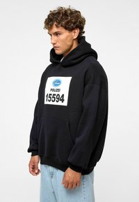 Sweatshirt à capuche noir avec un grand graphique blanc portant "POLIZEI 15594" sur le devant, poche sur le devant et poignets côtelés.