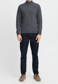 Maglione grigio scuro a maglia con collo alto e zip frontale, abbinato a jeans blu scuro e scarpe marrone chiaro.