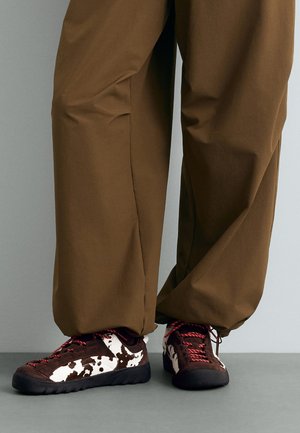 Pantalon large marron couvrant les chevilles, associé à des baskets noir et blanc à motif vache avec des lacets rouges sur un sol gris.