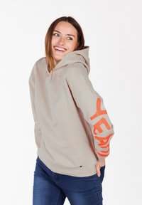 Felpa beige realizzata in materiale morbido con una grande grafica arancione "YEAH" sulla manica. Ha un cappuccio ampio e una vestibilità rilassata.