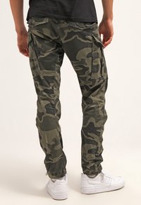 Pantalons cargo camouflage en vert foncé, dotés de plusieurs poches, d'une coupe décontractée et d'un tissu texturé. Portés avec des baskets blanches.