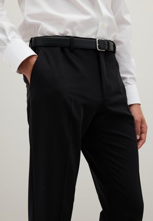 Schwarze, maßgeschneiderte Hose mit glatter Textur, ausgestattet mit Seitentaschen und einem schwarzen Ledergürtel mit einer silbernen Schnalle. Weiße Hemdmanschetten sichtbar.