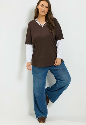 Kobieta z długimi brązowymi włosami, ubrana w brązowy krótki t-shirt z dekoltem w serek na białą koszulę z długim rękawem, niebieskie szerokie dżinsy i brązowe buty.