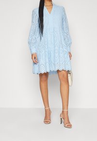 Ljusblå klänning med lång ärm, ögla detaljer, scallopad nederkant och lager-design. Matchad med naken klacksandaler och en liten handväska.