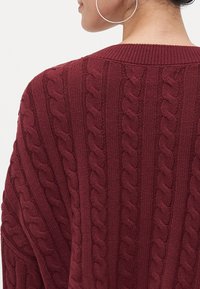 Burgundy knitted pullover featuring a cable knit pattern, ribbed collar, and loose fit, showcasing textured details on the fabric.