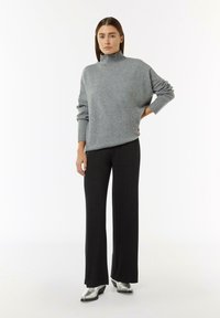 Pull en maille gris à col roulé avec des poignets côtelés, associé à un pantalon noir taille haute. Des chaussures argentées complètent la tenue.