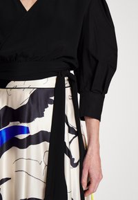 Gros plan d'une personne portant un haut cache-cœur noir à manches bouffantes et une jupe à motifs abstraits noirs et bleus.
