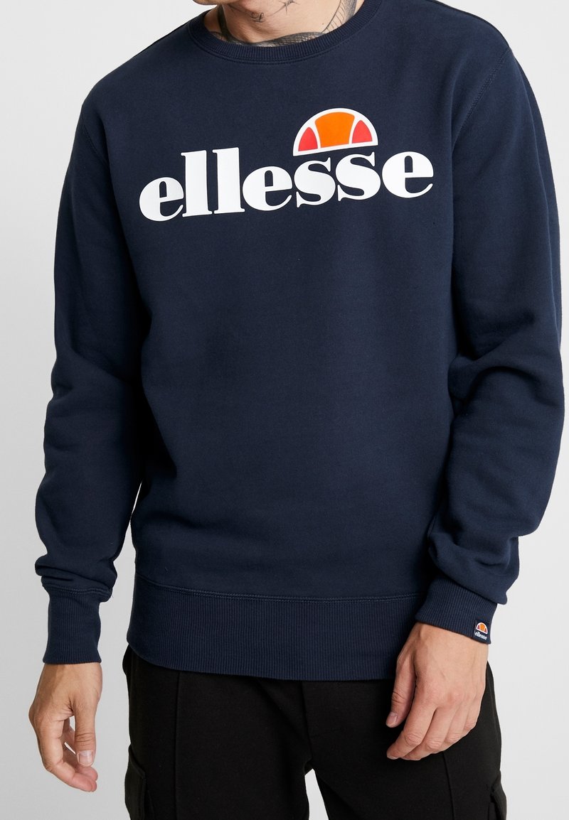 Homme portant un sweat-shirt bleu marine avec le logo blanc « ellesse » et un motif en demi-cercle orange sur la poitrine et le poignet, debout devant un fond uni.