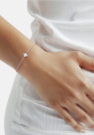Fijn zilveren armbandje met een kleine geometrische bedel, gedragen om een pols boven een witte, gestructureerde blouse.