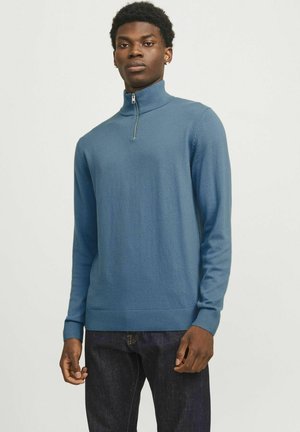 Suéter de cuello alto con cremallera azul, hecho de una tela suave, con mangas largas y puños acanalados, combinado con jeans oscuros. Textura lisa y diseño ajustado.