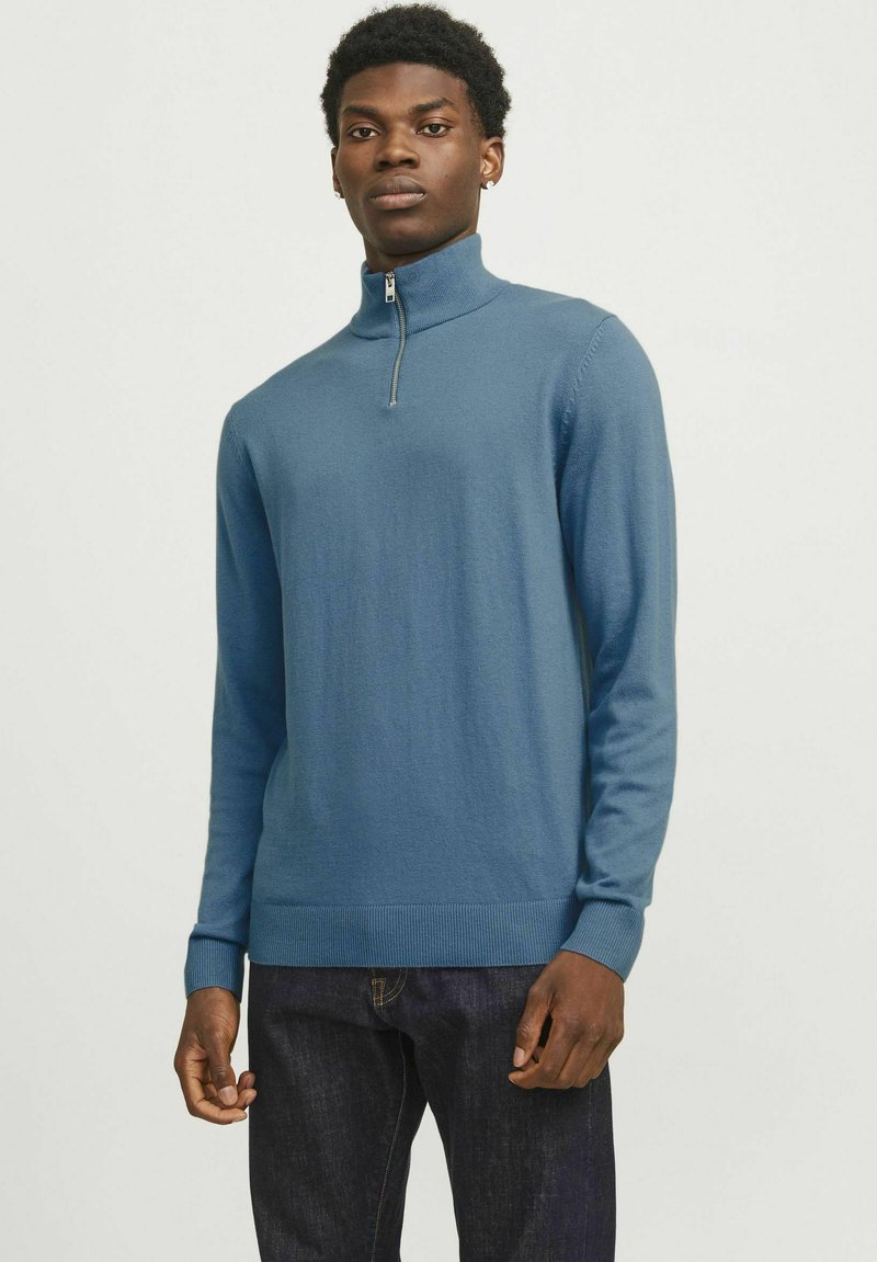 Suéter de cuello alto con cremallera azul, hecho de una tela suave, con mangas largas y puños acanalados, combinado con jeans oscuros. Textura lisa y diseño ajustado.