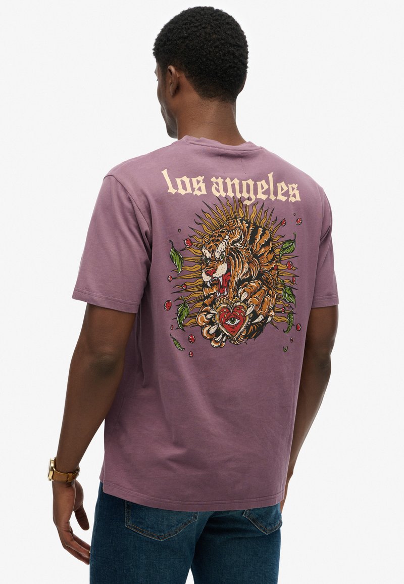 Man draagt een paarse t-shirt met een afbeelding van een brullende tijger die een hart met een oog vasthoudt, omringd door bladeren en de tekst "los angeles" op de rug.