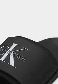 Κοντινό πλάνο μιας μαύρης σαγιονάρας slide με ανάγλυφο το λογότυπο Calvin Klein σε λευκό και μαύρο στο λουράκι.