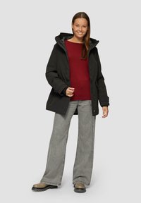Schwarze isolierte Jacke mit Kapuze, mit einem Reißverschluss und seitlichen Taschen. Getragen über einem bordeauxfarbenen Oberteil und kombiniert mit grauen weit geschnittenen Hosen.