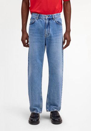 Mężczyzna w jasnoniebieskich prostych dżinsach, czarnych mokasynach i czerwonej koszuli, stojący na tle jednolitego białego tła.