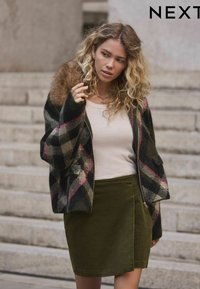 Kockás mintájú, fekete, zöld és rózsaszín kötött dzseki, műszőrme gallérral; egy testhezálló olajzöld szoknyával párosítva.