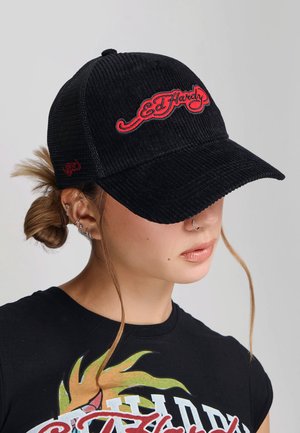 Młoda kobieta z wieloma kolczykami w uszach, nosząca czarną aksamitną czapkę Ed Hardy i czarną koszulkę z kolorowym nadrukiem.