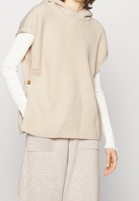 Top senza maniche in maglia beige con cappuccio e bottoni laterali, indossato sopra una camicia a maniche lunghe bianca. Texture morbida, design casual.