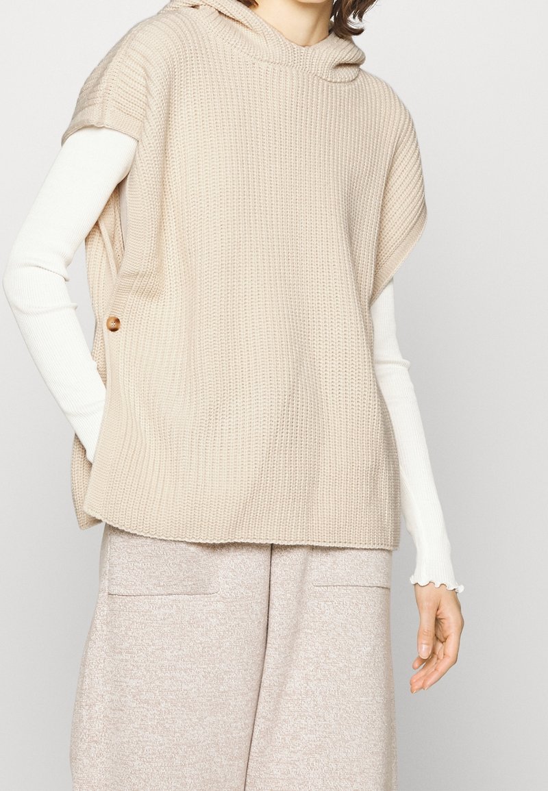 Top senza maniche in maglia beige con cappuccio e bottoni laterali, indossato sopra una camicia a maniche lunghe bianca. Texture morbida, design casual.